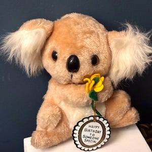 May include: A plush koala bear with tan fur and fluffy white ears, holding a yellow flower. The koala has a black nose and large, dark eyes. A tag reads "HAPPY BIRTHDAY TO SOMEONE SPECIAL."