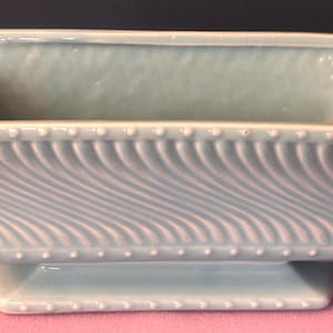 May include: A light blue ceramic planter with a rectangular shape and a pedestal base. The planter features a textured, swirled design on the sides and a beaded trim along the top edge. The planter measures approximately 8 inches long.