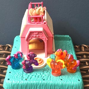 May include: A pink toy horse stable with a white roof and a light blue base. Several small, colorful toy horses are scattered around the stable. One horse is on the balcony. The horses are various colors, including blue, purple, orange, and pink.