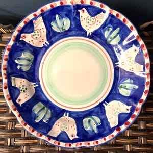 May include: A hand-painted ceramic plate with a blue rim and a white center. The plate features a repeating pattern of white birds with red and green accents, and green leaf-like shapes. The rim has red dots.