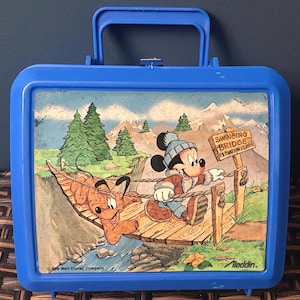 May include: Blue vintage Aladdin lunchbox featuring Mickey Mouse and Pluto crossing a wooden bridge. The illustration includes a sign that reads "Swinging Bridge Elevation 13,000 ft." The lunchbox has a blue handle and clasp.