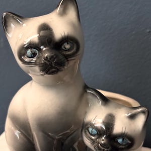 May include: A ceramic Siamese cat figurine with a smaller kitten. The cats have black markings on their faces, ears, and paws, with blue eyes. The larger cat is seated, and the smaller cat is nestled beside it.