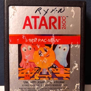 May include: A black Atari 2600 video game cartridge for Ms. Pac-Man. The silver label features the Atari logo, the game title, and a graphic of Ms. Pac-Man with pink and blue ghosts. The name Ryan is handwritten in black ink at the top of the label.