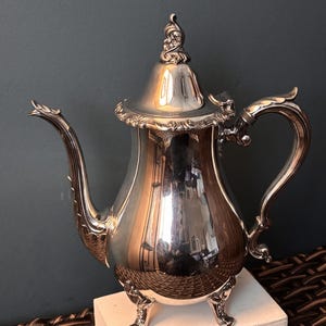 May include: A silver-colored teapot with ornate detailing. The teapot has a curved spout, a decorative handle, and sits on three small feet. The lid is topped with a decorative finial. The teapot is highly reflective, showing the surrounding environment.