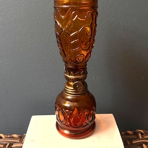 May include: An amber-colored glass oil lamp featuring a textured, embossed pattern on both the bulbous base and the cylindrical top section. A metallic brass-toned burner assembly with a circular adjustment knob sits between the two glass parts.