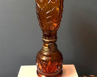 May include: An amber-colored glass oil lamp featuring a textured, embossed pattern on both the bulbous base and the cylindrical top section. A metallic brass-toned burner assembly with a circular adjustment knob sits between the two glass parts.