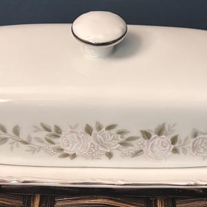 May include: A white porcelain butter dish with a lid and a matching base. The dish is decorated with a floral pattern of white roses and green leaves. The lid has a small, round knob. The edges are trimmed with a silver-colored band.