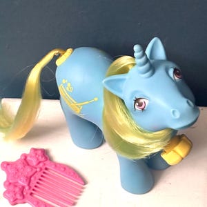 May include: A light blue unicorn toy with yellow hair and tail, and a yellow accessory on its front leg. A pink star-shaped comb is next to the unicorn. The unicorn has a horn and a musical instrument design on its side.