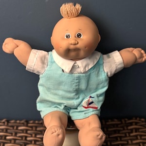 May include: A vintage Cabbage Patch Kid doll with a tan complexion, brown eyes, and a tuft of blonde hair. The doll wears a light blue corduroy romper with a white collar and a sailboat applique.