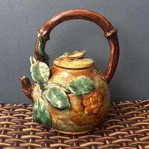 May include: A ceramic teapot with a brown, arched handle and a lid. The body is decorated with green leaves and a cluster of brown grapes. The teapot has a brown spout and is set against a dark background.