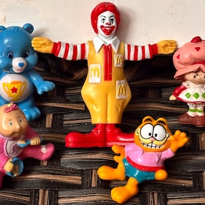 May include: A collection of vintage character figurines, including a blue Care Bear, Ronald McDonald, Strawberry Shortcake, a baby doll, and Garfield. The figures are brightly colored and made of plastic.