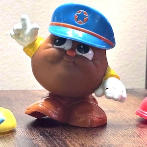 May include: A brown, anthropomorphic potato toy wearing a blue hat with a red and orange trim and a star emblem. The toy has white gloves, a yellow collar, and brown shoes. The toy is posed with one arm raised.