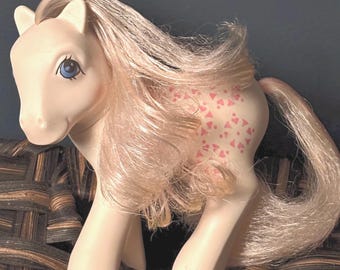 May include: A vintage toy pony in a standing position. The pony is primarily off-white with pink hair and a pink heart pattern on its side. It has blue eyes and is posed on a woven surface.
