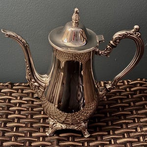 May include: A silver-colored coffee pot with ornate detailing. The pot features a curved spout, a decorative handle, and a lid with a finial. The body of the pot has a textured design. The pot is sitting on a woven surface.
