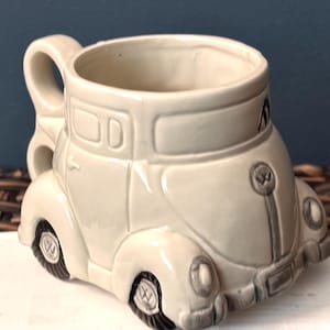 May include: A cream-colored ceramic mug shaped like a vintage car. The mug features details like headlights, wheels, and a handle. The car mug has a glossy finish and is designed to resemble a classic automobile.