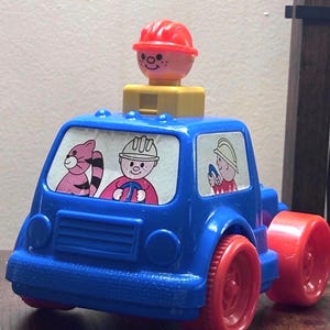 May include: A vintage blue toy truck with red wheels. The truck features cartoon illustrations in the windows and a smiling figure with a red hard hat on top. A classic children's toy.