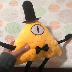 May include: A plush yellow triangle-shaped character with a black top hat, bow tie, and limbs. The character has a large, single eye and is a cartoon-like design. The plush toy is held by a person.