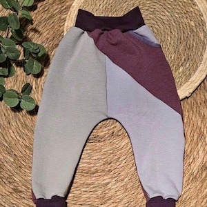 Könnte beinhalten: Babyhose mit Farbblöcken in Grau, Lila und Burgunder. Die Hose hat einen hohen Bund und Bündchen in einem dunkleren Lilaton. Das Design weist ein geometrisches Farbblockmuster auf. Die Hose ist aus weichem Stoff gefertigt.