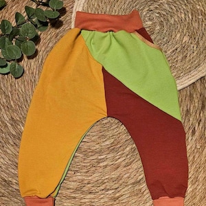 Op de afbeelding: Kleurrijke babybroek in een patchwork design. De broek heeft panelen van gele, groene en roodbruine stof, met oranje manchetten en tailleband. Het kledingstuk is gemaakt van zacht materiaal, geschikt voor baby's en peuters.