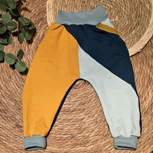 Könnte beinhalten: Babyhose mit Farbblöcken in Senfgelb, Marineblau und Hellgrau. Die Hose hat einen hohen, grauen Bund und passende Bündchen. Das Design zeichnet sich durch geometrische Farbblöcke aus und schafft einen modernen und verspielten Look für Babys und Kleinkinder.