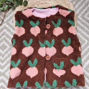 May include: Brown fleece vest with a pink lining and a pattern of pink and green beet-like shapes. The vest has two wooden buttons and a small pocket. The vest is designed for children.