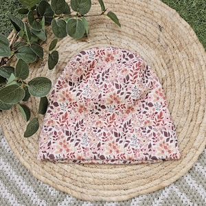 Puede incluir: Un gorro con estampado floral en tonos rosa, burdeos y crema. El gorro está hecho de una tela acanalada y está colocado sobre una estera de paja tejida. El fondo incluye follaje verde y una alfombra estampada.