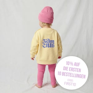 Op de afbeelding: Een kind draagt een lichtgele trui met blauwe tekst "5am club", gecombineerd met een roze legging en een roze muts. De afbeelding bevat een promotionele afbeelding met tekst.
