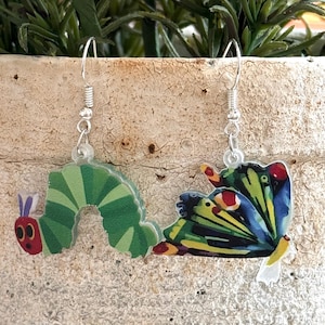 Puede incluir: Un par de pendientes colgantes con un diseño de oruga verde y mariposa colorida. La oruga tiene detalles rojos y morados. La mariposa presenta detalles azules, amarillos y rojos. Los pendientes cuelgan de ganchos plateados.