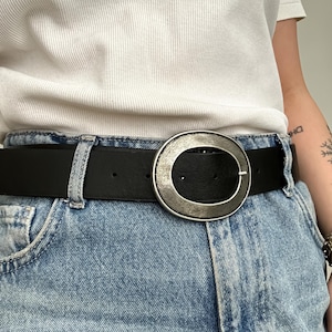 May include: Black leather belt with a large, silver-toned oval buckle. The belt is worn with light blue denim jeans. The buckle has a textured, antique finish. The belt is a classic accessory.