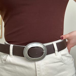 May include: A brown belt with a large, oval, silver-toned buckle. The belt is worn with cream-colored pants and a brown top. The buckle is the focal point, adding a stylish accent to the outfit.