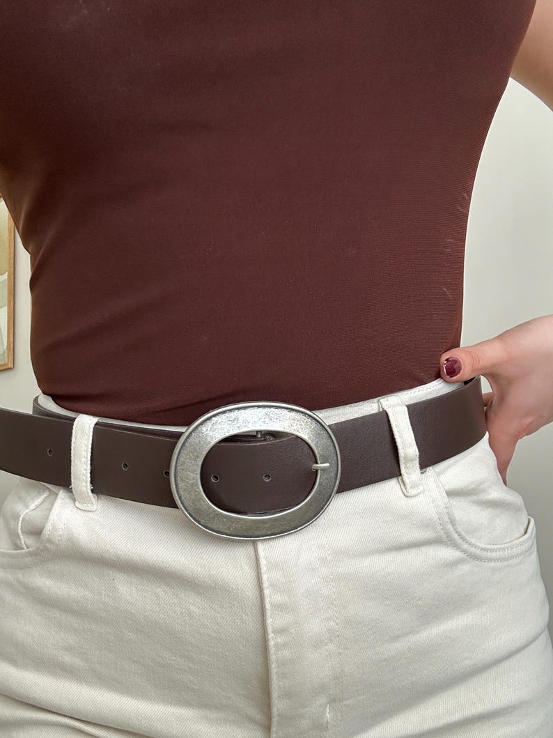 May include: A brown belt with a large, oval, silver-toned buckle. The belt is worn with cream-colored pants and a brown top. The buckle is the focal point, adding a stylish accent to the outfit.