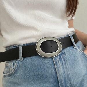 May include: Black leather belt with a large, oval silver buckle. The buckle has a textured, metallic finish. The belt is worn with light blue denim jeans and a white ribbed top.