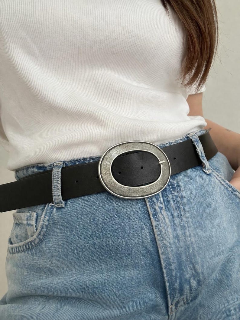 May include: Black leather belt with a large, oval silver buckle. The buckle has a textured, metallic finish. The belt is worn with light blue denim jeans and a white ribbed top.