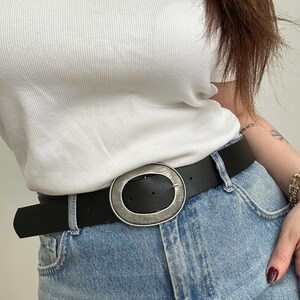 May include: Black belt with a large, oval, silver-toned buckle. The belt is worn with light blue denim jeans and a white ribbed top. The buckle has a distressed finish, adding a vintage look to the accessory.