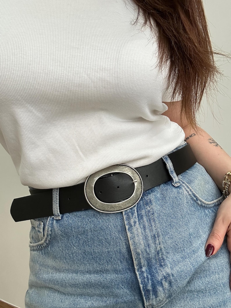 May include: Black belt with a large, oval, silver-toned buckle. The belt is worn with light blue denim jeans and a white ribbed top. The buckle has a distressed finish, adding a vintage look to the accessory.