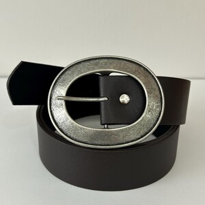 May include: A dark brown belt with a large, round, silver-toned buckle. The buckle has a textured, antique finish. The belt is made of a smooth, solid material and is rolled up. The buckle has a small, silver-toned stud.