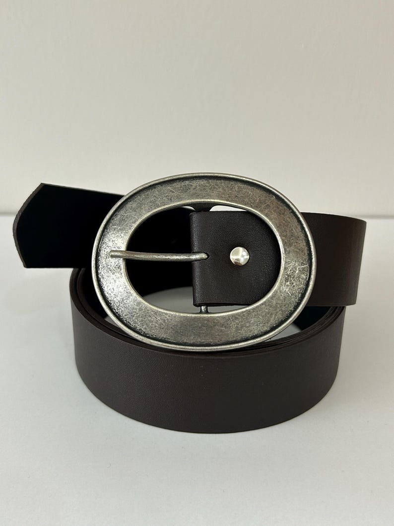 May include: A dark brown belt with a large, round, silver-toned buckle. The buckle has a textured, antique finish. The belt is made of a smooth, solid material and is rolled up. The buckle has a small, silver-toned stud.