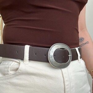 May include: A brown leather belt with a large, silver-colored circular buckle. The belt is worn with white jeans and a brown top. The buckle is a prominent feature, adding a stylish accent to the outfit.