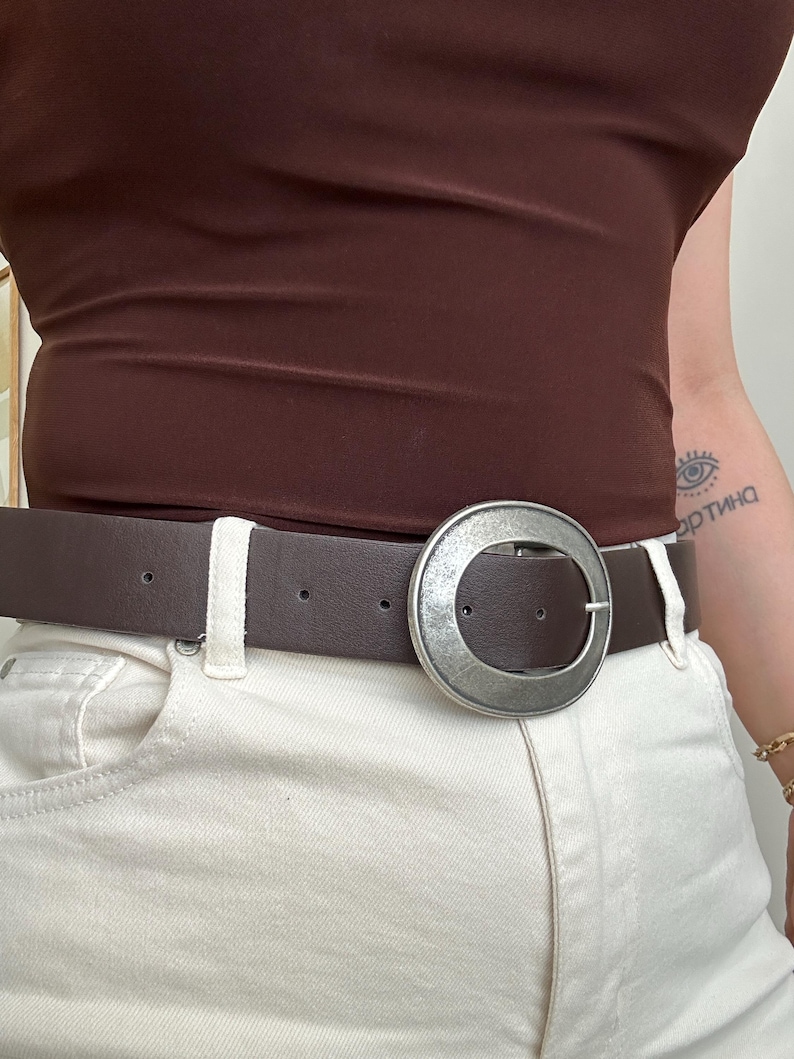 May include: A brown leather belt with a large, silver-colored circular buckle. The belt is worn with white jeans and a brown top. The buckle is a prominent feature, adding a stylish accent to the outfit.