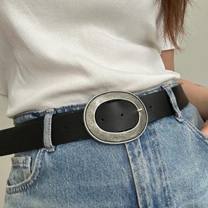 May include: A black leather belt with a large, silver-toned oval buckle. The belt is worn with light blue denim jeans. The buckle has a textured, metallic finish. The belt is approximately 1.5 inches wide.
