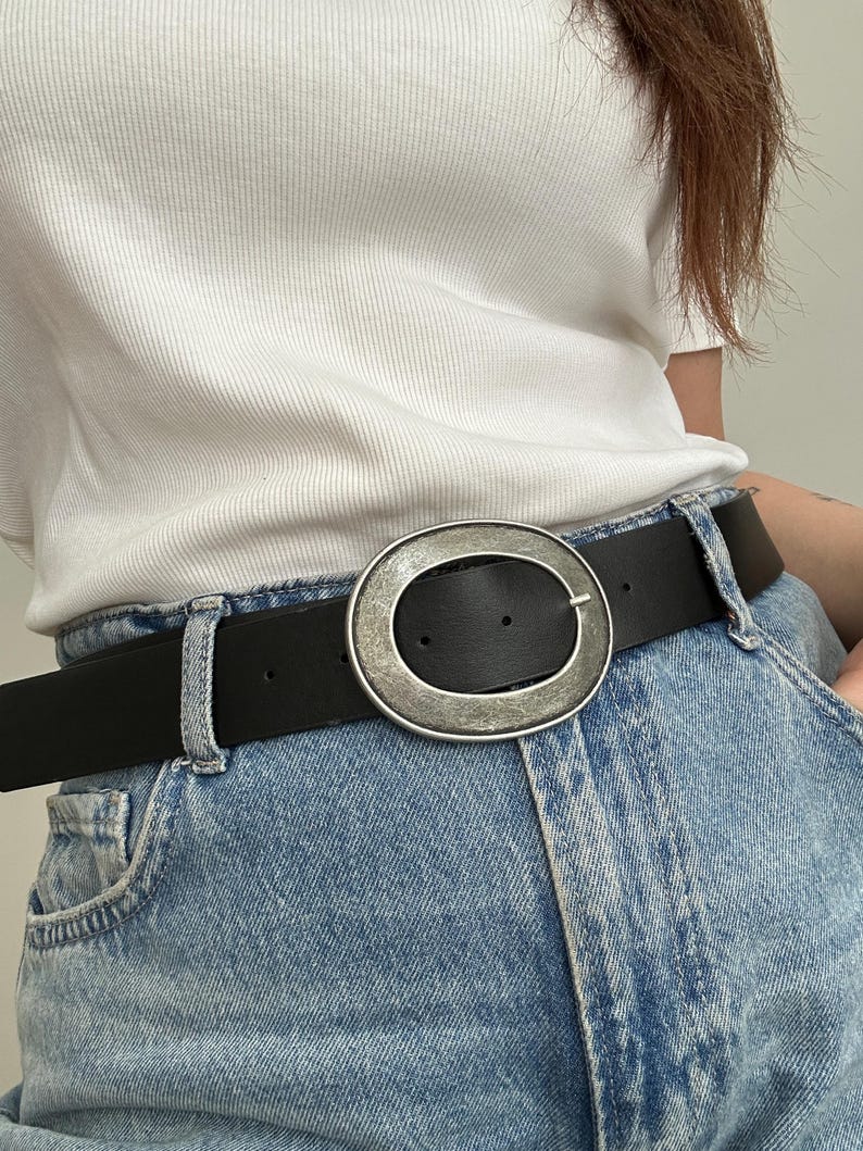 May include: A black leather belt with a large, silver-toned oval buckle. The belt is worn with light blue denim jeans. The buckle has a textured, metallic finish. The belt is approximately 1.5 inches wide.