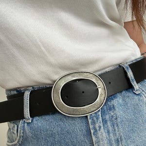 May include: Black leather belt with a large, oval, silver-toned buckle. The belt is worn with blue jeans and a white t-shirt. The buckle has a textured appearance.
