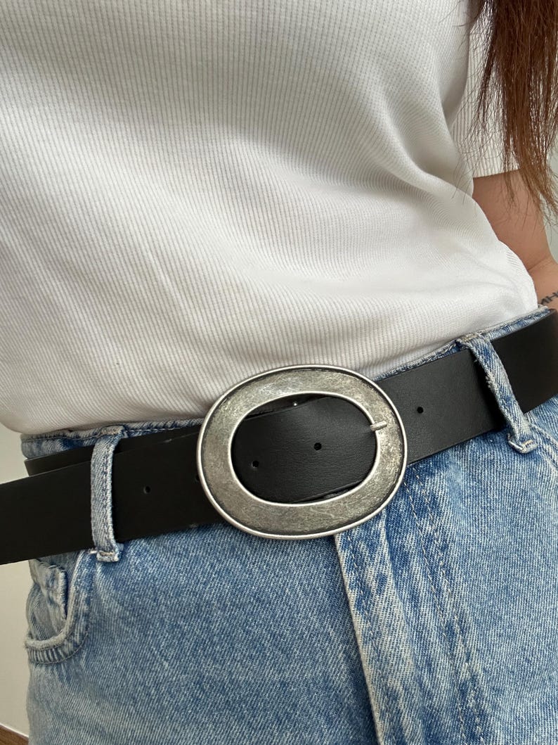 May include: Black leather belt with a large, oval, silver-toned buckle. The belt is worn with blue jeans and a white t-shirt. The buckle has a textured appearance.