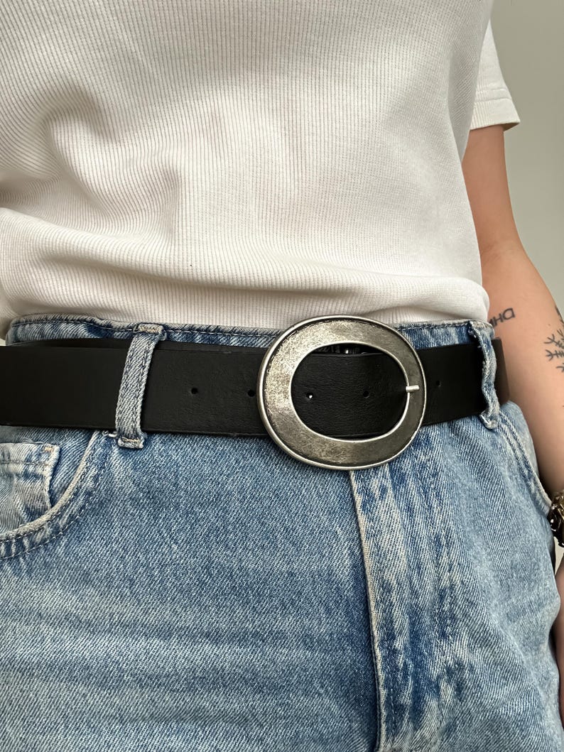 May include: Black leather belt with a large, silver-toned oval buckle. The belt is worn with light blue denim jeans. The buckle has a textured, antique finish. The belt is a classic accessory.