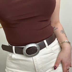 May include: A brown leather belt with a large, oval, silver-toned buckle. The belt is worn with white jeans and a brown top. The buckle is the focal point, adding a touch of style to the outfit.