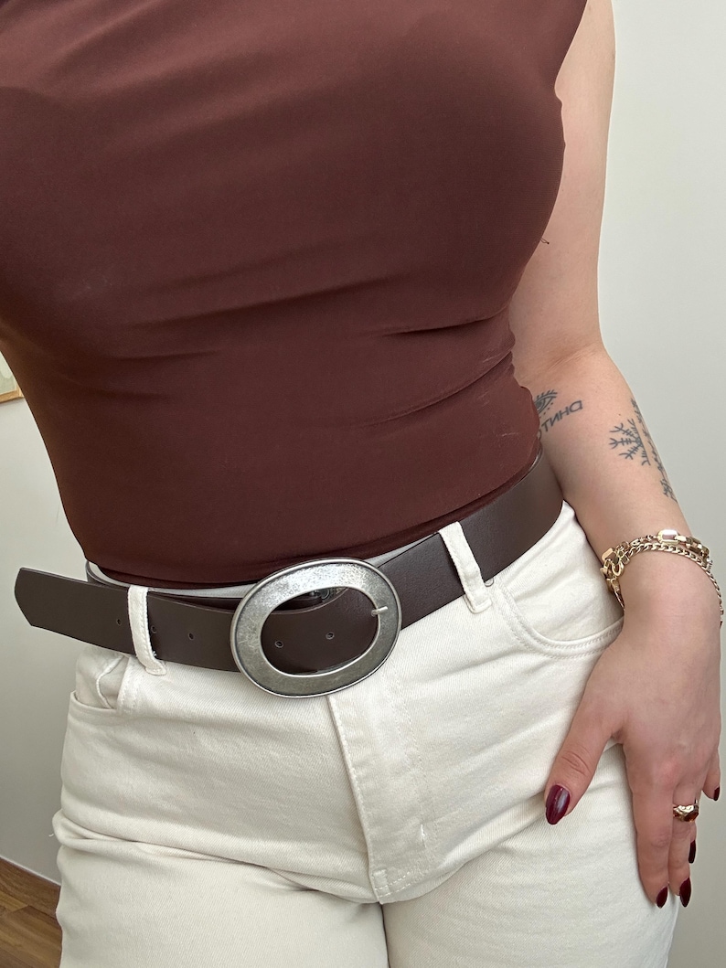 May include: A brown leather belt with a large, oval, silver-toned buckle. The belt is worn with white jeans and a brown top. The buckle is the focal point, adding a touch of style to the outfit.