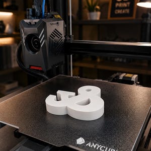 Może przedstawiać: Biały symbol ampersand wydrukowany w technologii 3D, spoczywający na teksturowanym czarnym stole roboczym drukarki 3D Anycubic. Głowica drukująca znajduje się nad płytą roboczą, kt&oacute;ra w prawym dolnym rogu posiada białe logo Anycubic.