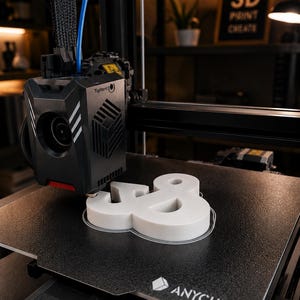 Może przedstawiać: Drukarka 3D Anycubic z czarną głowicą drukującą i teksturowaną płytą roboczą. Drukarka tworzy biały, tr&oacute;jwymiarowy symbol ampersand. Na głowicy drukującej widoczny jest napis Tighten oraz logo firmy Anycubic na płycie roboczej.