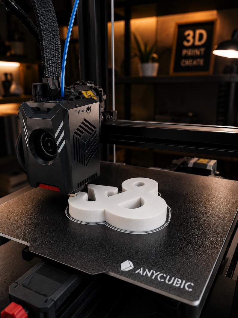Może przedstawiać: Drukarka 3D Anycubic z czarną głowicą drukującą i teksturowaną płytą roboczą. Drukarka tworzy biały, tr&oacute;jwymiarowy symbol ampersand. Na głowicy drukującej widoczny jest napis Tighten oraz logo firmy Anycubic na płycie roboczej.
