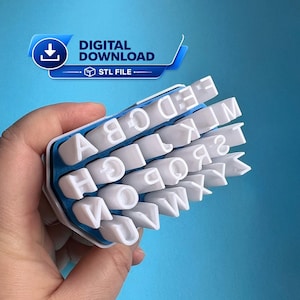 May include: A white 3D printed alphabet stamp set featuring raised capital letters arranged on a blue base. The letters A through Z are clearly visible in a grid-like formation. A blue banner at the top displays the text DIGITAL DOWNLOAD STL FILE.